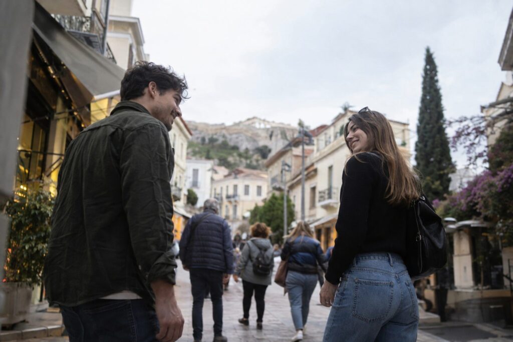 Δύο άτομα σε καφέ χαμογελούν και ανταλλάσσουν βλέμματα, συνδέοντας με ευγενικό φλερτ χωρίς εφαρμογές.