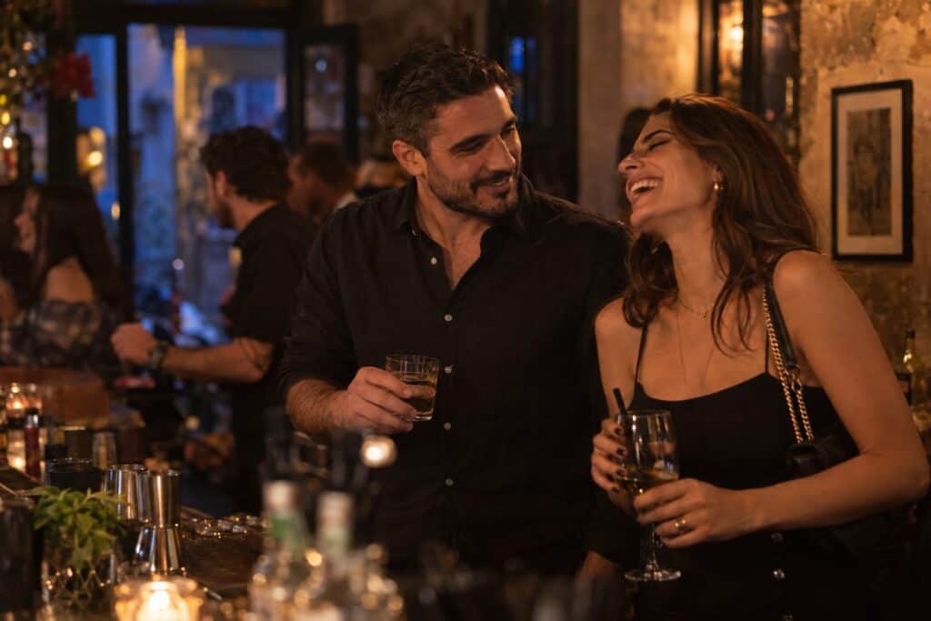 Έλληνας άντρας στα 30 μιλάει με μια γυναίκα στο μπαρ, εκείνη γελάει, ζεστό φως, φλερτ και μήνυμα