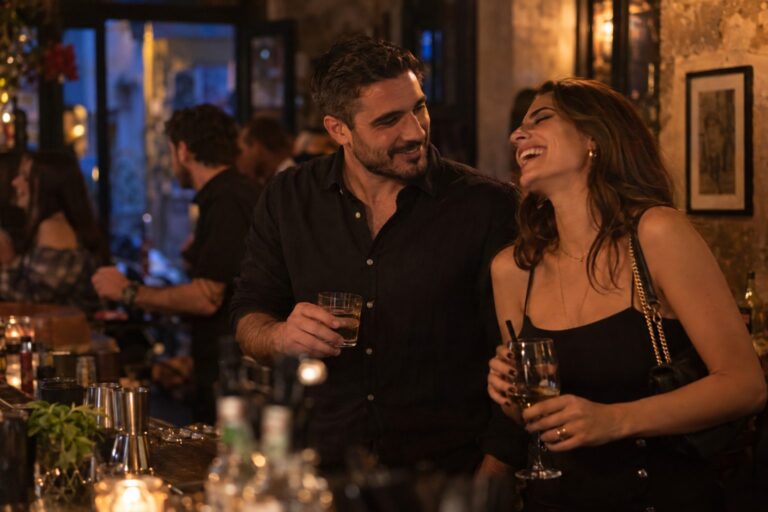 Έλληνας άντρας στα 30 μιλάει με μια γυναίκα στο μπαρ, εκείνη γελάει, ζεστό φως, φλερτ και μήνυμα