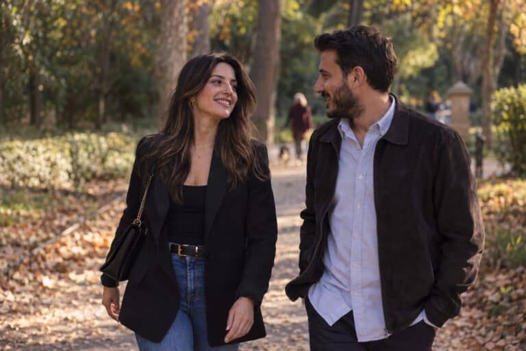 Ζευγάρι Ελλήνων περπατά στον Εθνικό Κήπο της Αθήνας, φθινοπωρινά φύλλα κάτω, χαμογελώντας και συζητώντας στις αρχές σχέσης.