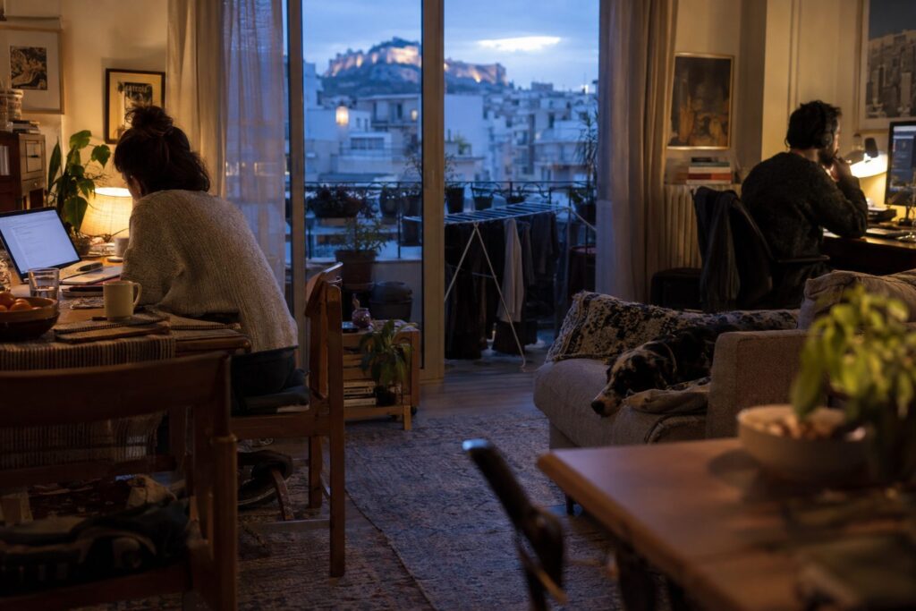 Ζευγάρι σε σπίτι-γραφείο με δύο φορητούς υπολογιστές, σημειώσεις και ζώνη ήσυχης εργασίας.