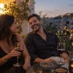 Έλληνας άνδρας γύρω στα 40 γελάει με τη σύντροφό του σε μπαλκόνι, συζητούν ερωτήσεις, δύο ποτήρια κρασί, φώτα πόλης πίσω.