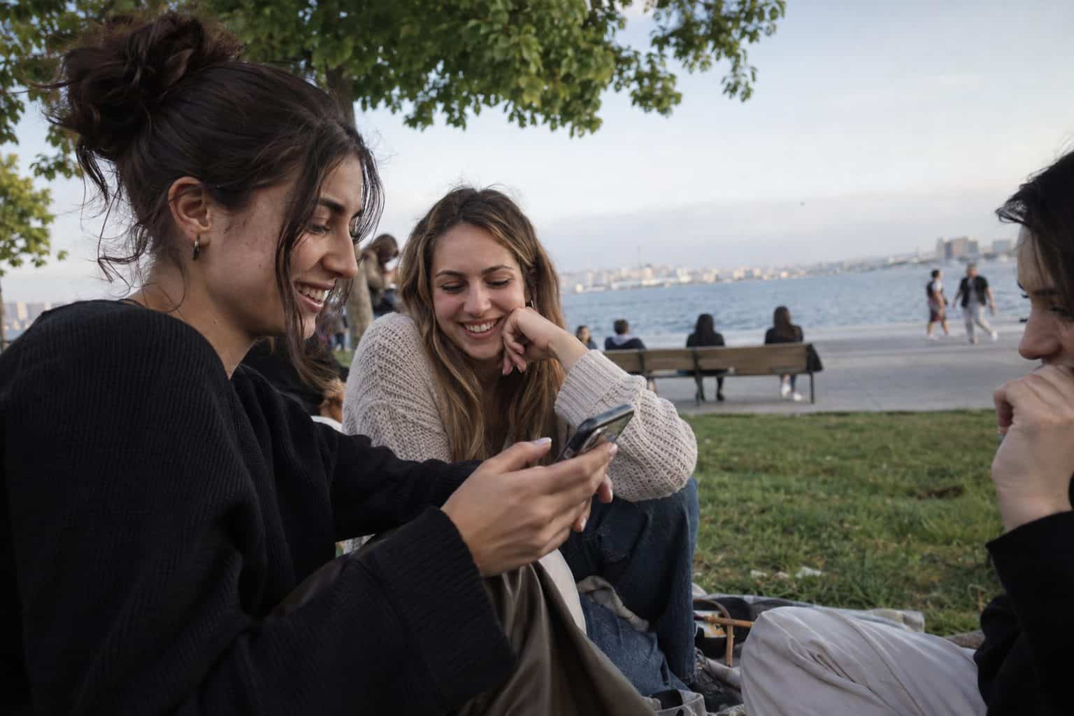 Ομάδα φίλων γύρω από τραπέζι βλέπει ομαδικό chat σε κινητά και σχολιάζει το δεύτερο ραντεβού σήμερα.