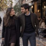 Ζευγάρι σε οδό με δέντρα και πεσμένα φύλλα, ντυμένοι φθινοπωρινά για ραντεβού, πίσω τραπεζάκια καφέ.