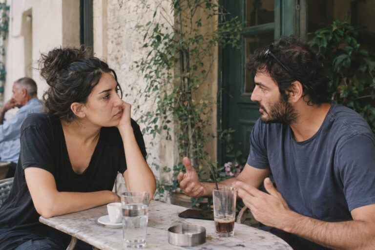 Ζευγάρι σε σχέση κάθεται σε καναπέ, κουβεντιάζει ήρεμα για πολιτικά θέματα, μπροστά τους χαρτιά, δείχνοντας όρια.