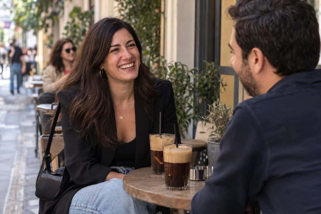 Γυναίκα μέσης ηλικίας γελά απέναντι σε πρώτο καφέ ραντεβού, δύο παγωμένα καφέδες σε εξωτερικό τραπέζι στον πεζόδρομο Κουκάκι.