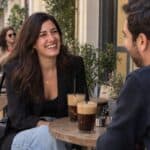 Γυναίκα μέσης ηλικίας γελά απέναντι σε πρώτο καφέ ραντεβού, δύο παγωμένα καφέδες σε εξωτερικό τραπέζι στον πεζόδρομο Κουκάκι.
