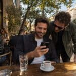 Έλληνας άντρας σε καφενείο της Κυψέλης χαμογελά βλέποντας το τηλέφωνο· φίλος κοντά του το παρατηρεί, σκηνή φλερτ με emoji.