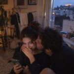 Ζευγάρι χαμογελά μπροστά σε κινητό με εικονίδια κοινωνικών δικτύων, συνδέοντας το θέμα Soft ή Hard Launch σχέσης.