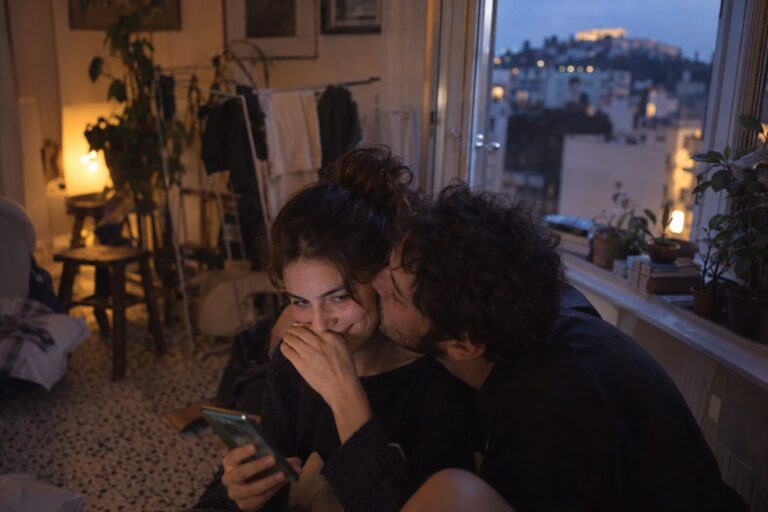 Ζευγάρι χαμογελά μπροστά σε κινητό με εικονίδια κοινωνικών δικτύων, συνδέοντας το θέμα Soft ή Hard Launch σχέσης.